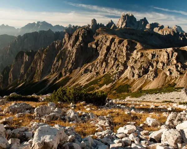Skaliste szczyty Dolomitów o wschodzie słońca, z kamienistym zboczem i suchą trawą na pierwszym planie.