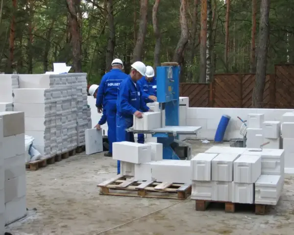 Budowa domu z bloczków betonowych. Pracownicy w niebieskich kombinezonach i białych kaskach przygotowują materiał. Widać stosy bloczków, piłę do cięcia i palety.