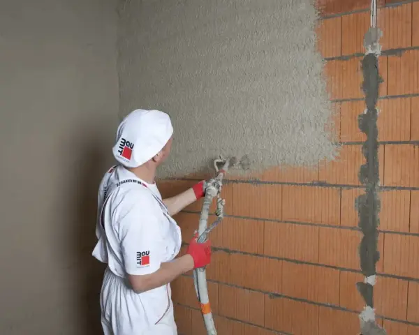 Pracownik w białym stroju nakłada tynki cementowo wapienne na ścianę z pustaków ceramicznych za pomocą agregatu tynkarskiego.