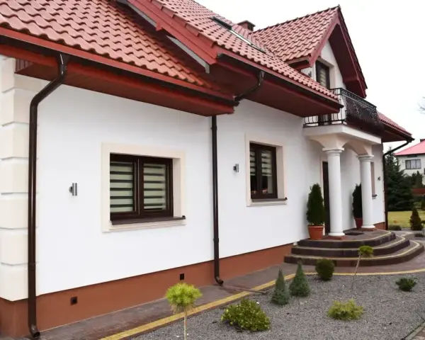 Biały dom z brązowymi cokołami, czerwonym dachem i ozdobnymi schodami.