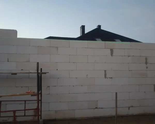 Budowa domu w toku. Ściany z bloczków betonowych czekają na tynkowanie. W tle widać dach i kominy.
