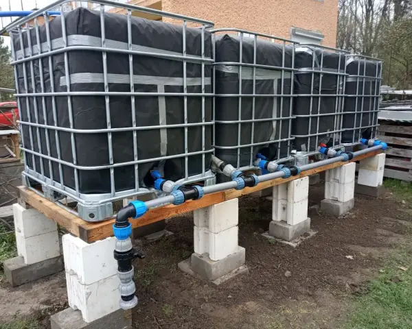 System zbiorników IBC, każdy z czarną osłoną, połączony rurami. Wygląda jak prowizoryczna instalacja do gromadzenia deszczówki, może nawet do nawadniania ogrodu, jak w przypadku systemu mauser.