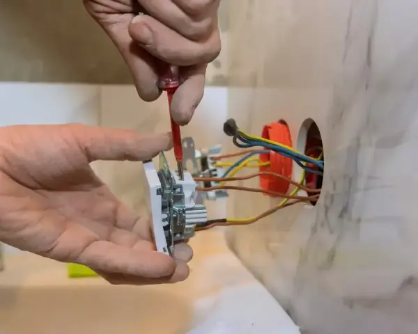 Instalator pokazuje, jak podłączyć gniazdko, przykręcając je do ściany. Widać przewody elektryczne.