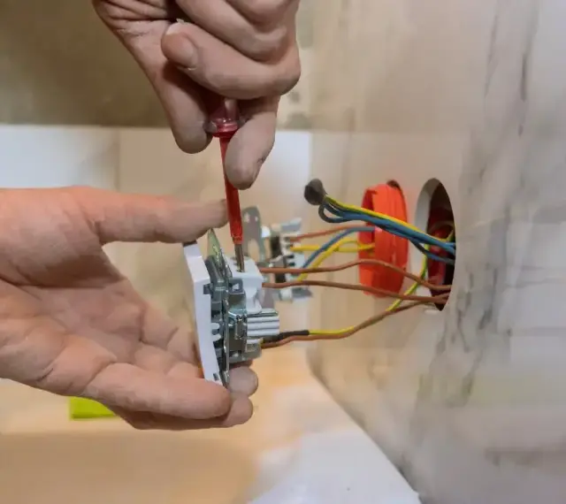 Instalator pokazuje, jak podłączyć gniazdko, przykręcając je do ściany. Widać przewody elektryczne.