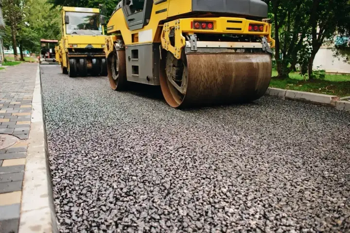 Walec drogowy ugniata świeży asfalt, tworząc gładką nawierzchnię. W tle widać kolejny walec i drzewa.