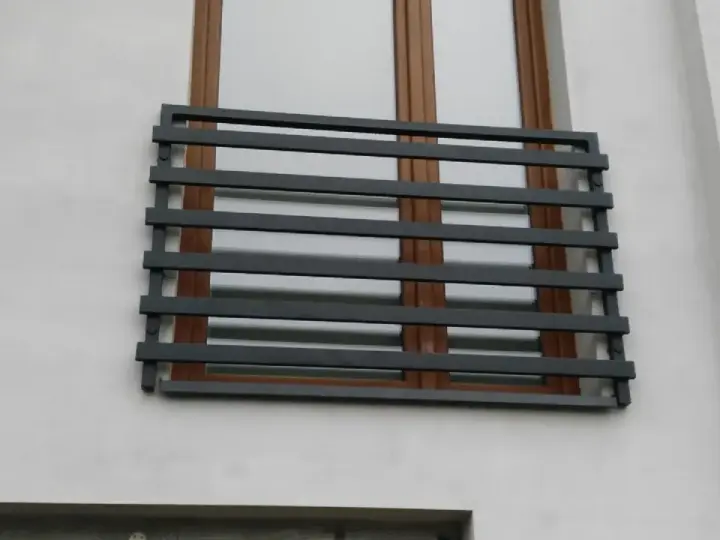 Nowoczesne balkony francuskie z ciemnoszarymi, poziomymi barierkami na tle białej ściany i brązowych okien.