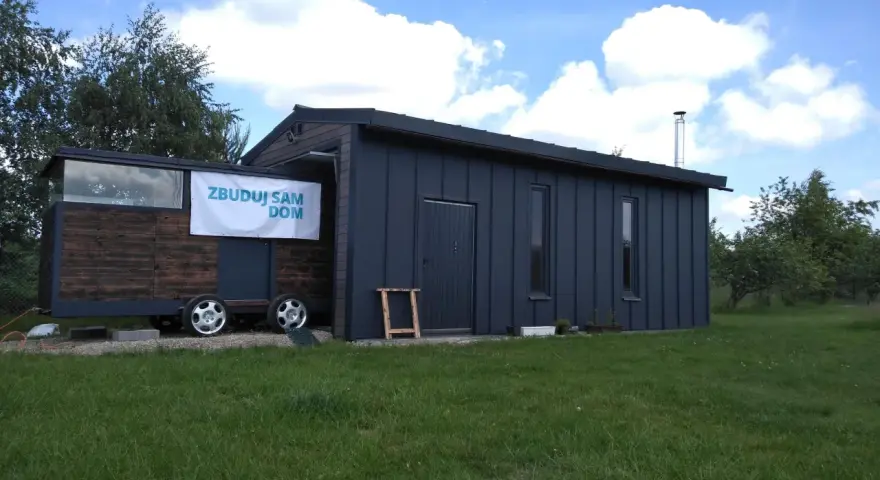 Mobilny garaż bez pozwolenia, z banerem "Zbuduj sam dom", stoi na trawie.