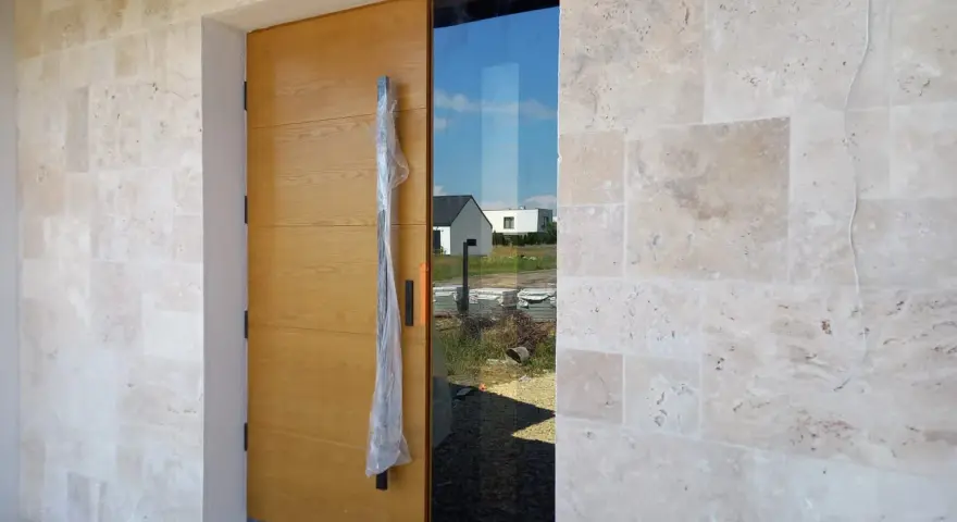 Elegancka fasada z trawertynu otacza nowoczesne, drewniane drzwi z bocznym przeszkleniem.