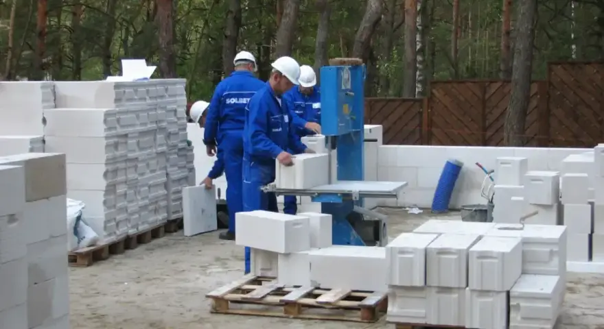 Budowa domu z bloczków betonowych. Pracownicy w niebieskich kombinezonach i białych kaskach przygotowują materiał. Widać stosy bloczków, piłę do cięcia i palety.