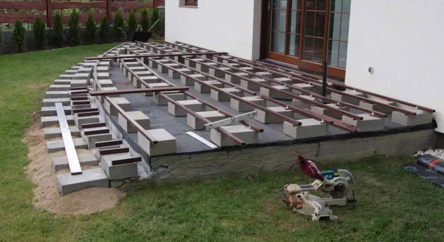 Budowa tarasu kompozytowego: betonowe bloczki podtrzymują legary, tworząc solidną konstrukcję. Widać narzędzia budowlane i materiały.