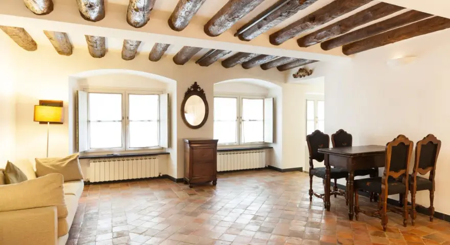 Stary salon z belkami stropowymi i podłogą z terakoty. Kanapa, stół z krzesłami i komoda tworzą przytulną atmosferę.