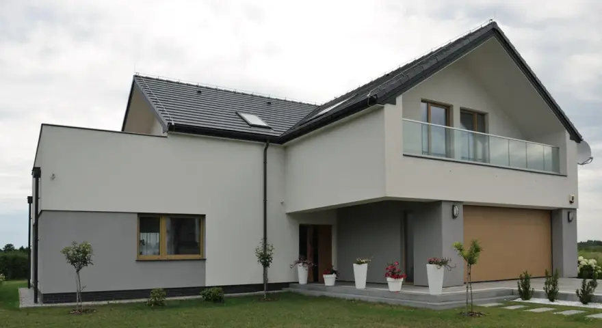 Nowoczesna elewacja budynku z szarymi i białymi tynkami, dużym oknem, balkonem i drewnianą bramą garażową.