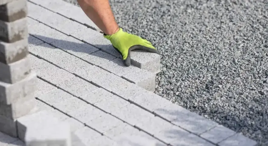 Ręka w zielonej rękawicy układa kostkę brukową na podsypce cementowo piaskowej.