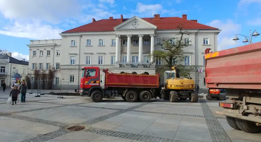Czerwona wywrotka i żółta koparka na placu przed budynkiem z kolumnami.