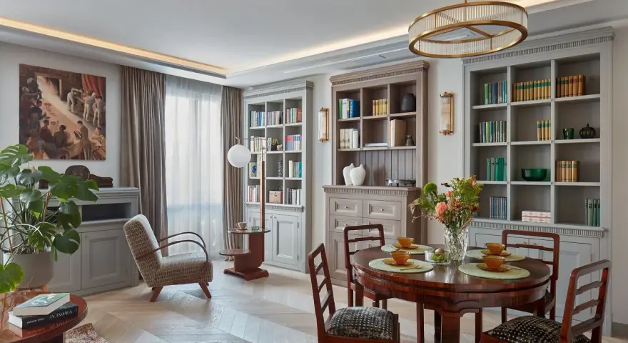 Przytulne pomieszczenie z jadalnią i biblioteczką. Okrągły stół z krzesłami, regały pełne książek, obraz na ścianie i wygodny fotel tworzą ciepłą atmosferę.
