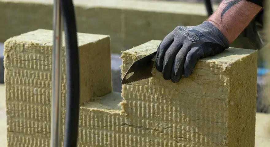 Ręka w rękawicy tnie piłą wełnę skalną 10. Materiał izolacyjny jest cięty na budowie.