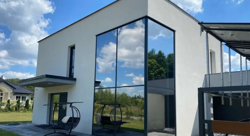 Nowoczesny dom z imponującym, narożnym oknem weneckim, w którym odbija się błękitne niebo z chmurami i zieleń drzew.
