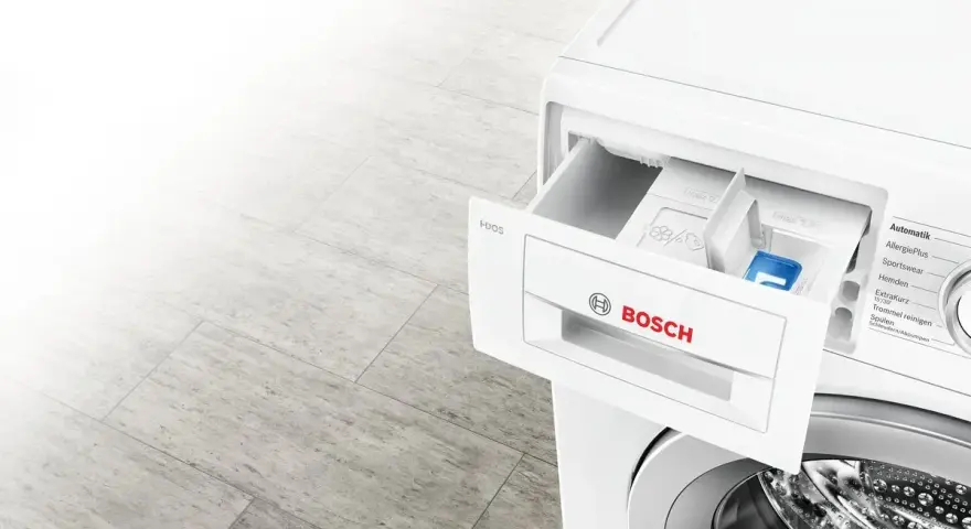Szara podłoga i biała pralka Bosch z wysuniętą szufladą na detergenty. Dowiedz się, ile wody zużywa pralka.