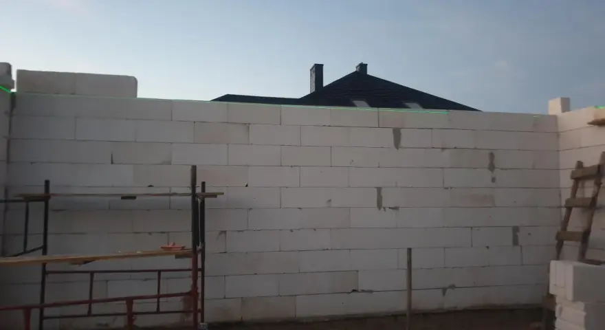 Budowa domu w toku. Ściany z bloczków betonowych czekają na tynkowanie. W tle widać dach i kominy.