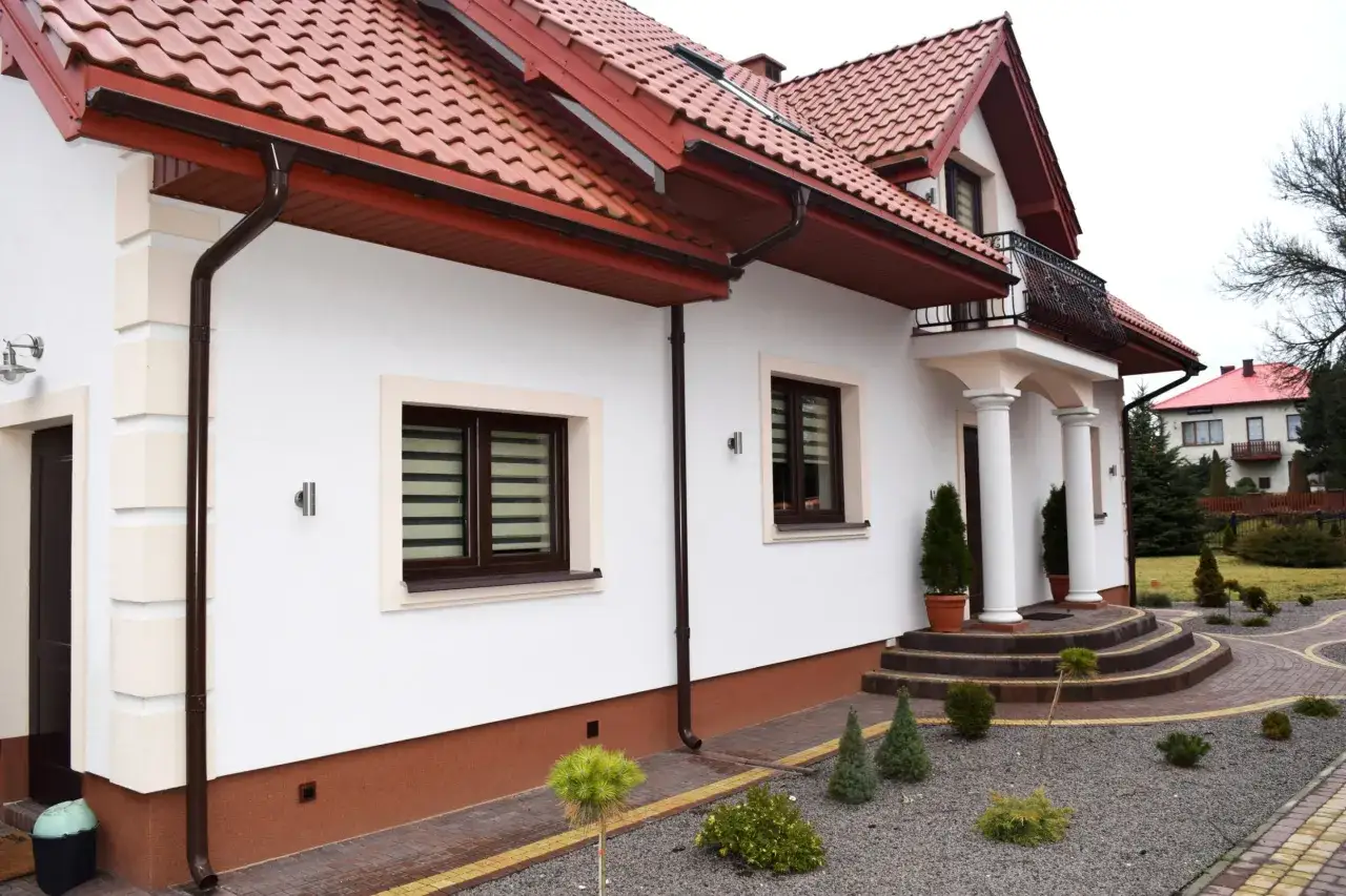 Nowoczesny dom z białym tynkiem, brązowym dachem i ozdobnym cokołem na elewacji. Wokół zadbany ogród z kamieniami i iglakami.