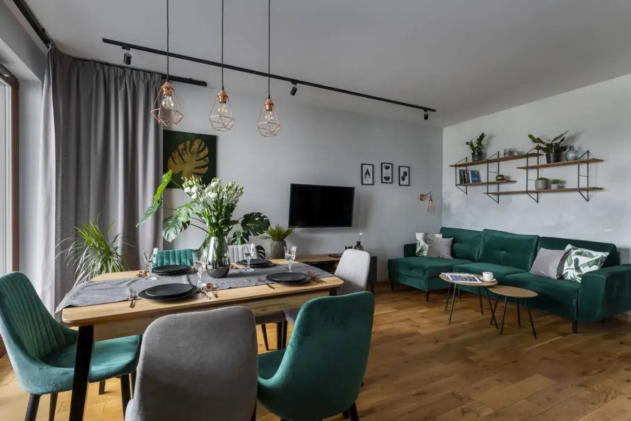 Nowoczesny salon z jadalnią. Szmaragdowa sofa i krzesła, drewniany stół, szare zasłony. Zastanawiasz się, jaki kolor pasuje do beżu? Zieleń i miedź świetnie się komponują!