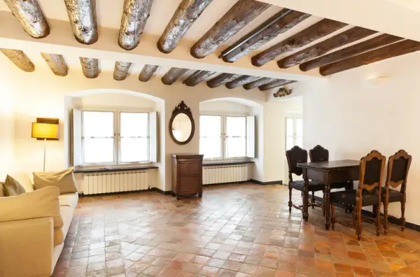Stary salon z belkami stropowymi i podłogą z terakoty. Kanapa, stół z krzesłami i komoda tworzą przytulną atmosferę.